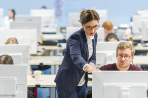 Eine Professorin erklärt einem jungen Mann etwas am Computermonitor. Sie sind in einem Klassenraum, in dem noch mehr Menschen an einem Computer sitzen.