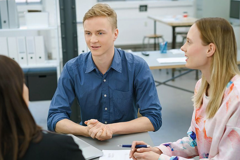 Ein junger Mann bespricht sich mit zwei jungen Kolleginnen, die mit ihm am Tisch sitzen.