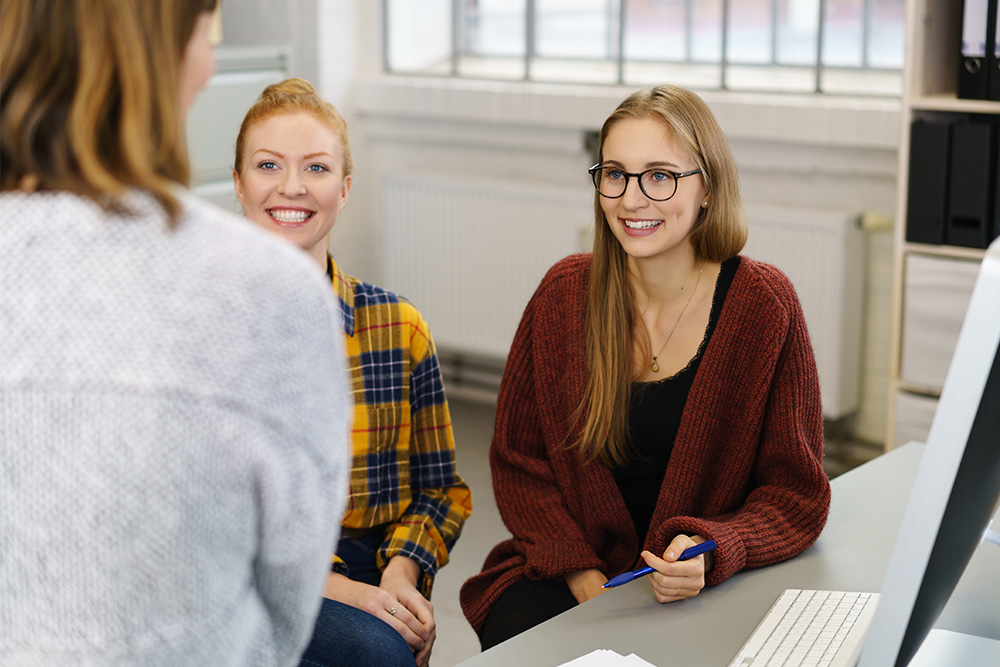 Zwei Auszubildende sitzen gemeinsam an einem Tisch und unterhalten sich mit einer Person, die gerade steht.