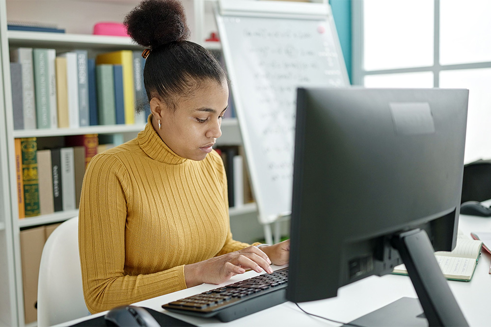 Eine junge Frau, die vor einem Bücherregal sitzt, tippt etwas in den Computer ein.