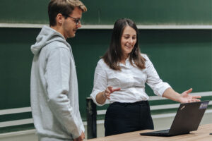 Eine junge Professorin erklärt ihrem Werkstudenten gerade etwas an ihrem Laptop.