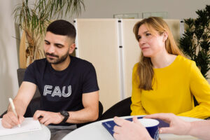 Ein junger Mann und eine junge Frau sitzen an einem Tisch. Der junge Mann notiert sich etwas. Die junge Frau hört jemanden zu, der nicht im Bild zu sehen ist.