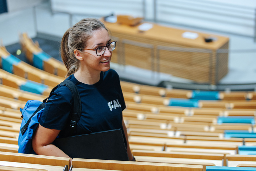 Eine junge Frau mit FAU-T-Shirt steht in einem Hörsaal in der oberen Sitzreihe.