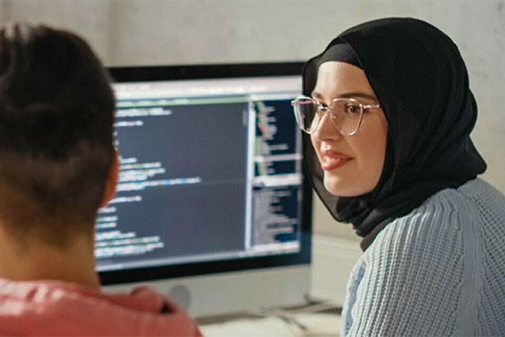 Eine junge Frau mit Kopftuch sieht zu ihrem Kollegen hinüber. Beide sitzen vor einem Monitor, auf dem eine Programmiersprache zu erkennen ist.